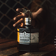A grimy hand reaches through a chain link fence to grab a bottle of Sexton Irish Whiskey.